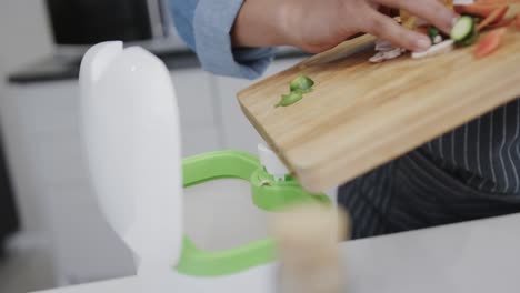 Hands-of-biracial-woman-in-apron-composting-vegetable-waste-in-kitchen,-slow-motion