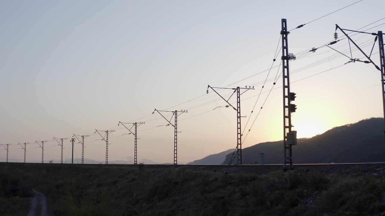 silueta de ferrocarril vacío con líneas eléctricas de alto voltaje y sol escondido detrás de una colina, al atardecer