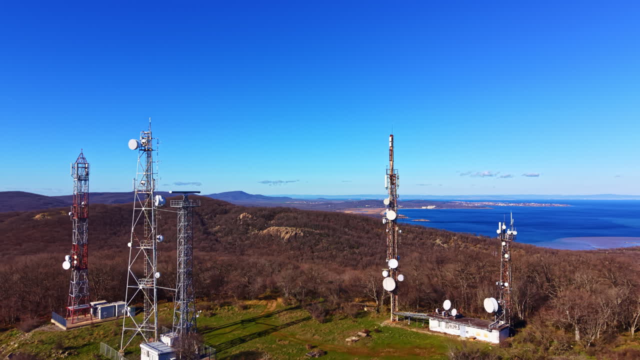 View of communication towers on a hill with blue sky and distant mountains during daytime near a lake