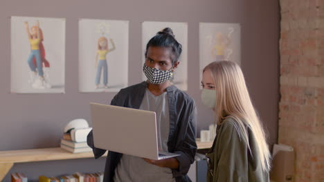 two multiethnic colleagues with face masks discussing a new project using laptop computer in an animation studio