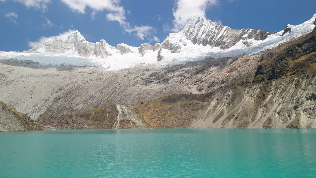 A stunning aerial tilt up shot of turquoise glacial lake beneath the majestic, snow-covered Cordillera Blanca mountains along Peru’s iconic Santa Cruz trek