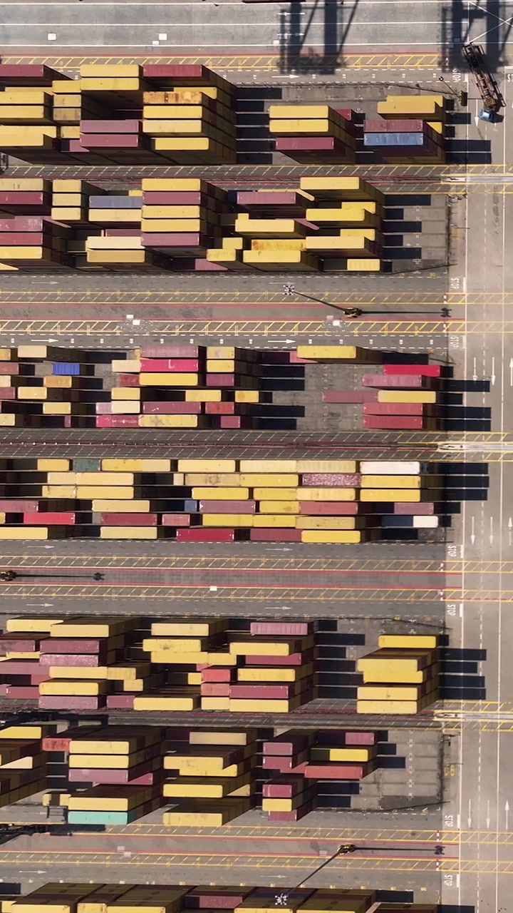 Aerial view of a busy container terminal showing numerous shipping containers arranged in rows. Cargo trucks transport goods within the terminal while a container ship is docked nearby.