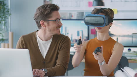 female architect wearing vr headset in office working with male colleague at desk with laptop