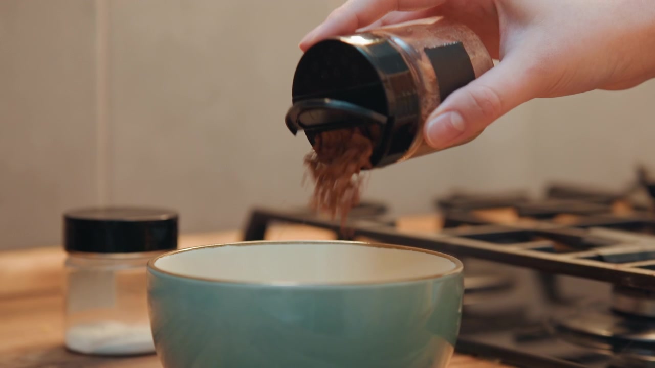 Versare spezie nella ciotola in modo naturale. Mani che versano con cura spezie aromatiche in una ciotola di ceramica vicina. Cascata di spezie mentre le mani inclinano il contenitore di ceramica sopra un piatto turchese in un ambiente da cucina.