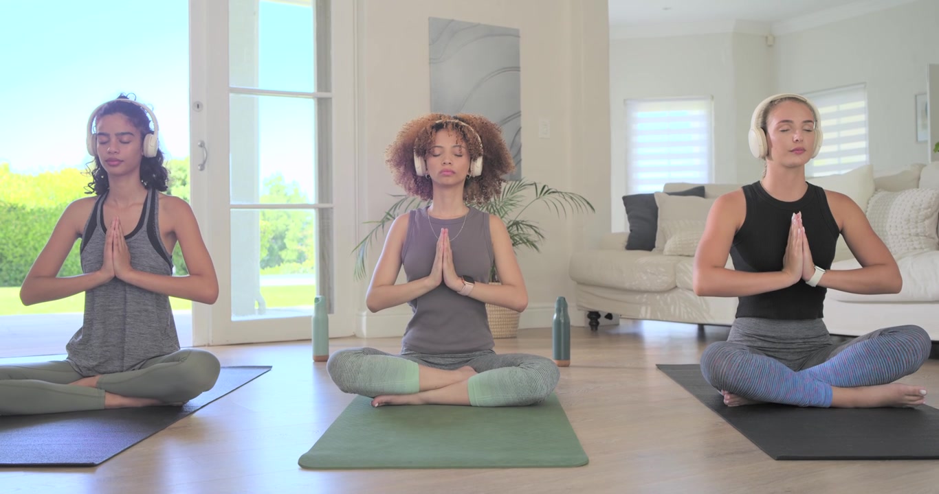 Gruppo eterogeneo di donne con cuffie su tappetini da yoga a casa, in posizione Anjali, che si calmano e poi aprono gli occhi.