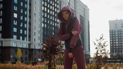 trainer in hoodie and vest stretches side body under bright sun in crisp morning breeze on urban pavement framed by residential towers and autumn landscaping during outdoor workout routine