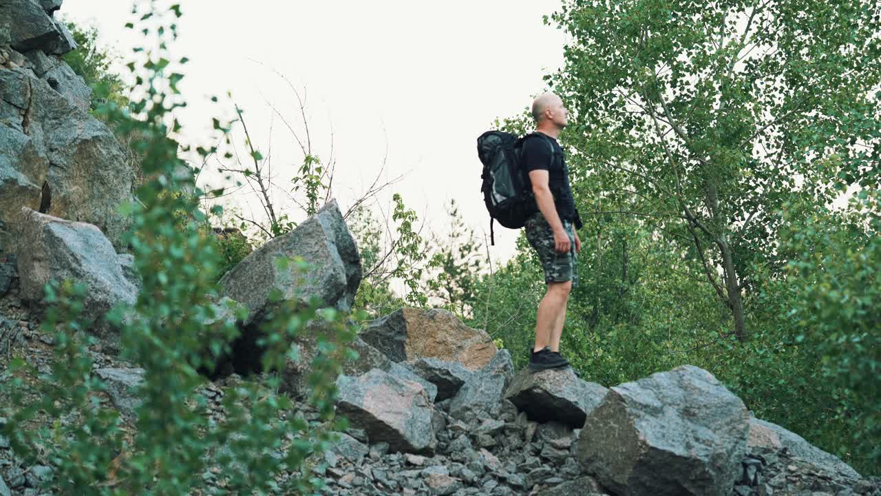 tourist in dark shorts and a t-shirt with a backpack on his shoulders is standing on the rocks and looking at the environment around himself on the background of green trees