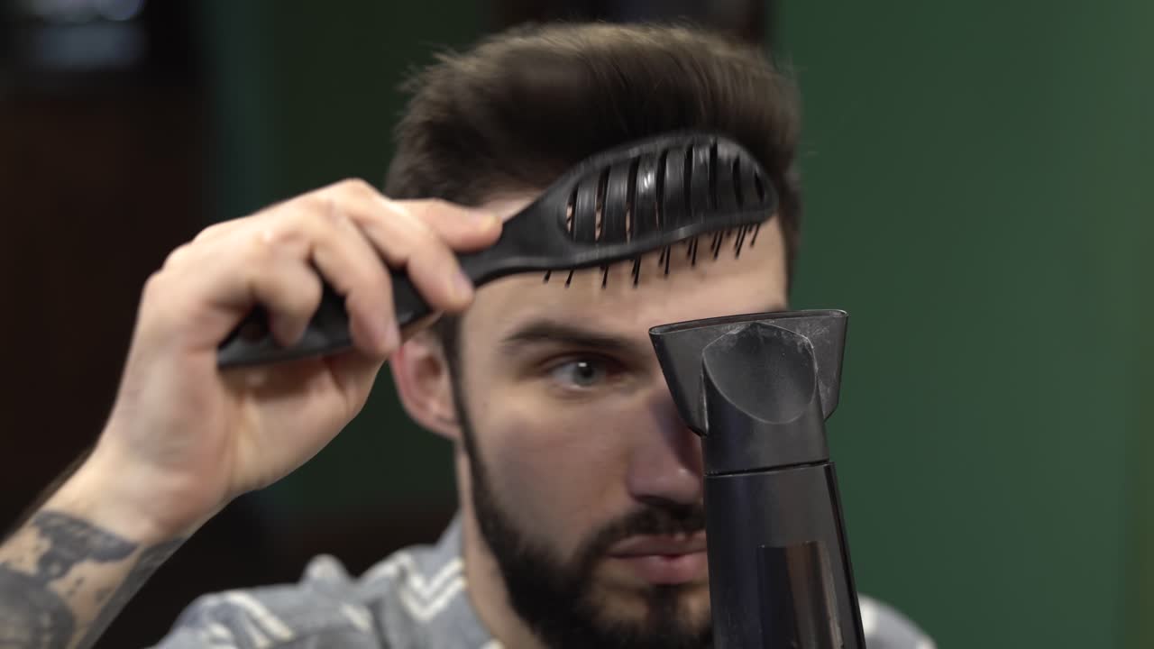 Young handsome bearded man is combing his hair with comb and hairdryer.