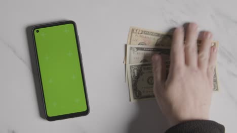 Overhead-Währungsaufnahme-Einer-Hand,-Die-US-Dollar-Scheine-Neben-Einem-Green-Screen-Handy-Ergreift