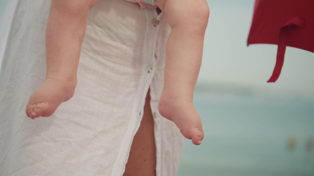 Riprese cinematografiche delicate di una madre che tiene in braccio un bambino vicino alla spiaggia, con mare calmo e ombrellone rosso sullo sfondo.