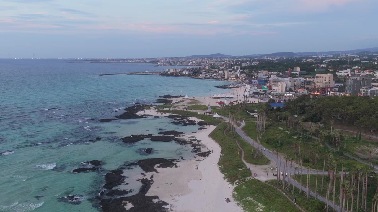 Inquadratura orizzontale con carrellata in avanti verso sinistra lungo la spiaggia di Hyeopjae, isola di Jeju, Corea del Sud, che mostra spiaggia sabbiosa, acqua turchese, rocce vulcaniche, costa e villaggio costiero.
