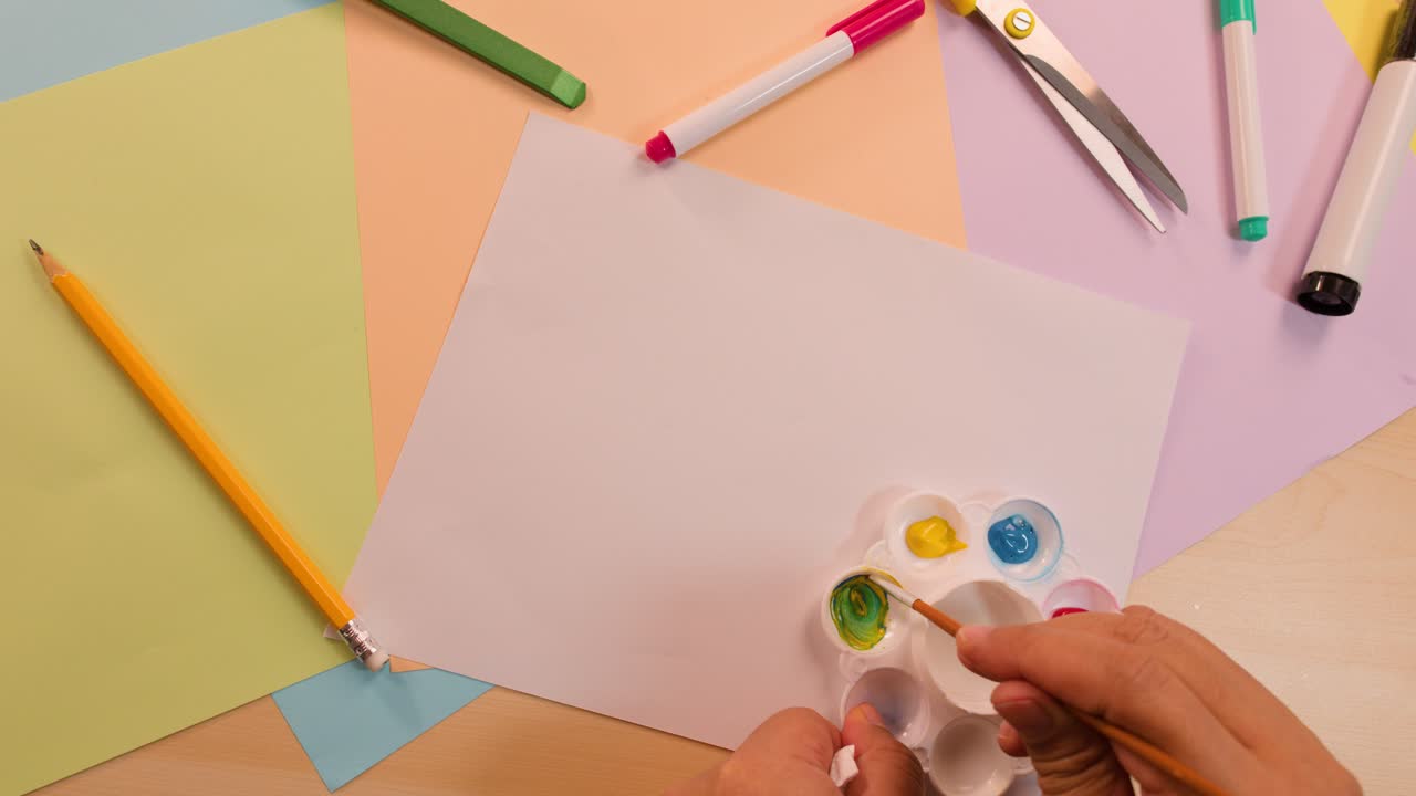 Hand mixes yellow and blue paint on palette above blank paper, colorful desk supplies visible