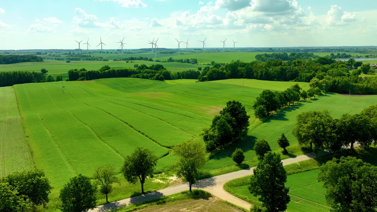 Expansive green fields with wind turbines and a tree-lined road under a blue sky with clouds