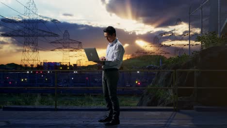 full body side view of an asian male professional worker standing with his laptop near high voltage tower, industrial facility, typing on his laptop's keybaord with meditation