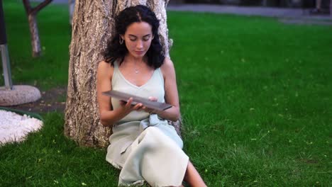 Woman-in-dress-comes-to-sit-below-tree-at-the-park,-she-opens-laptop,-put-it-on-her-knees-and-started-typing