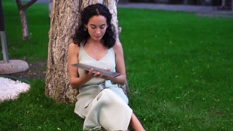 woman in dress comes to sit below tree at the park, she opens laptop, put it on her knees and started typing