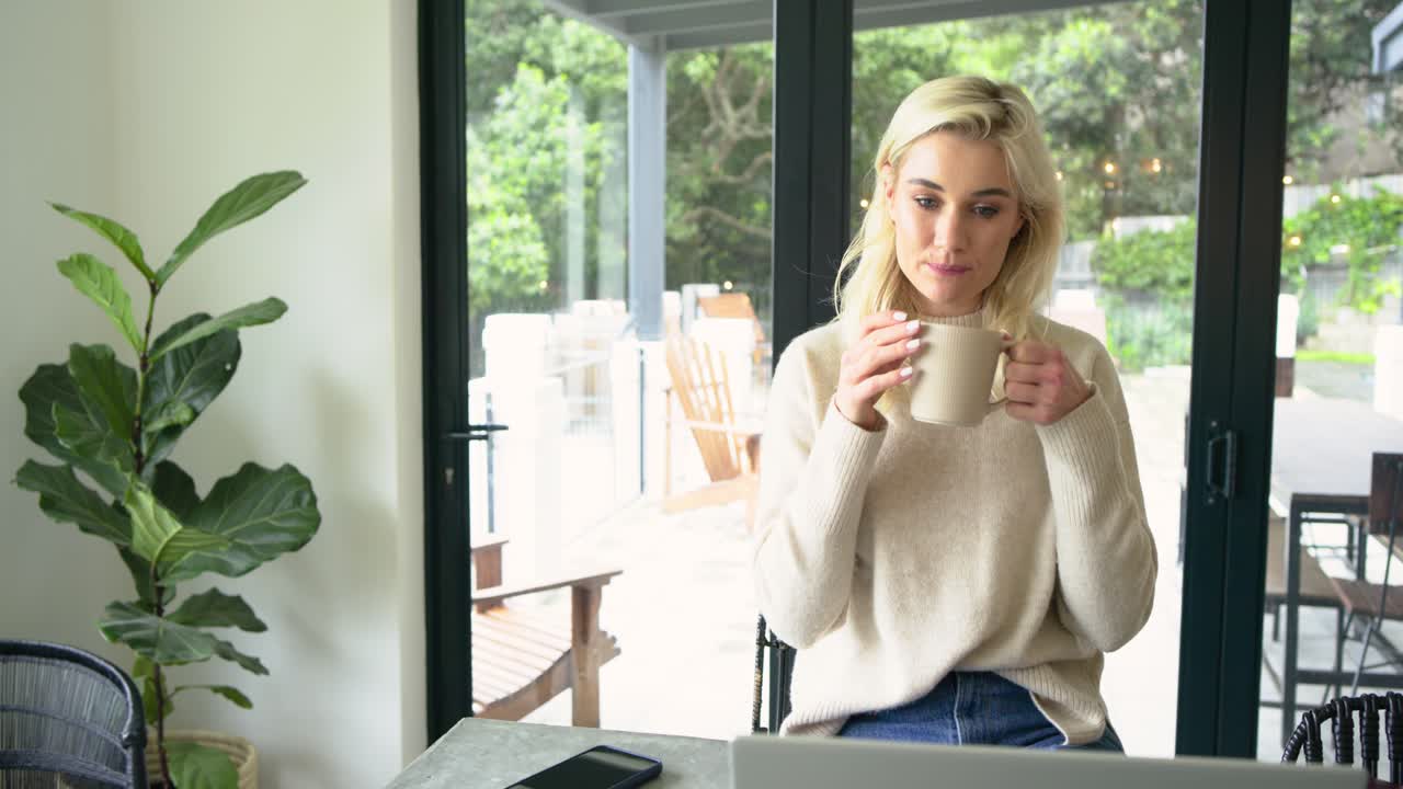 Reviewing laptop screen, female worker sipping coffee mug, typing for work at stone tabletop