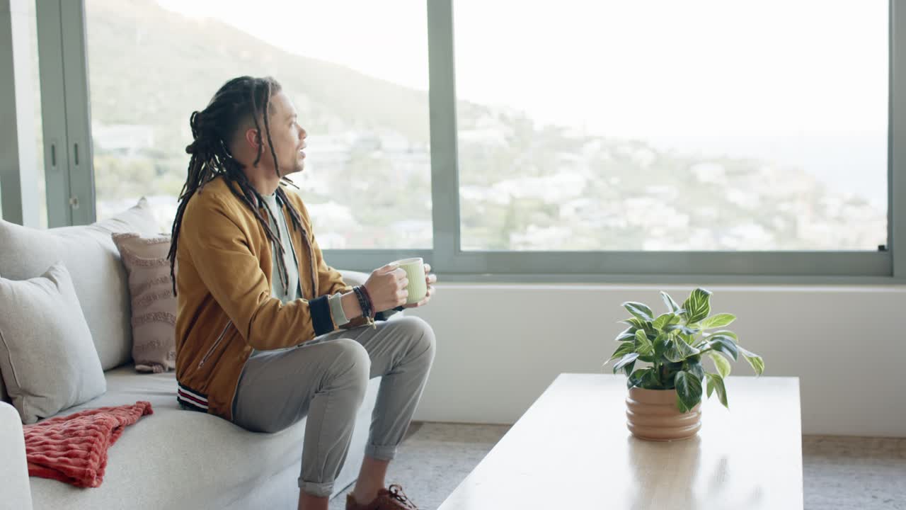 Mid adult man raising mug on sofa, sipping, gazing out window at hillside view, copy space