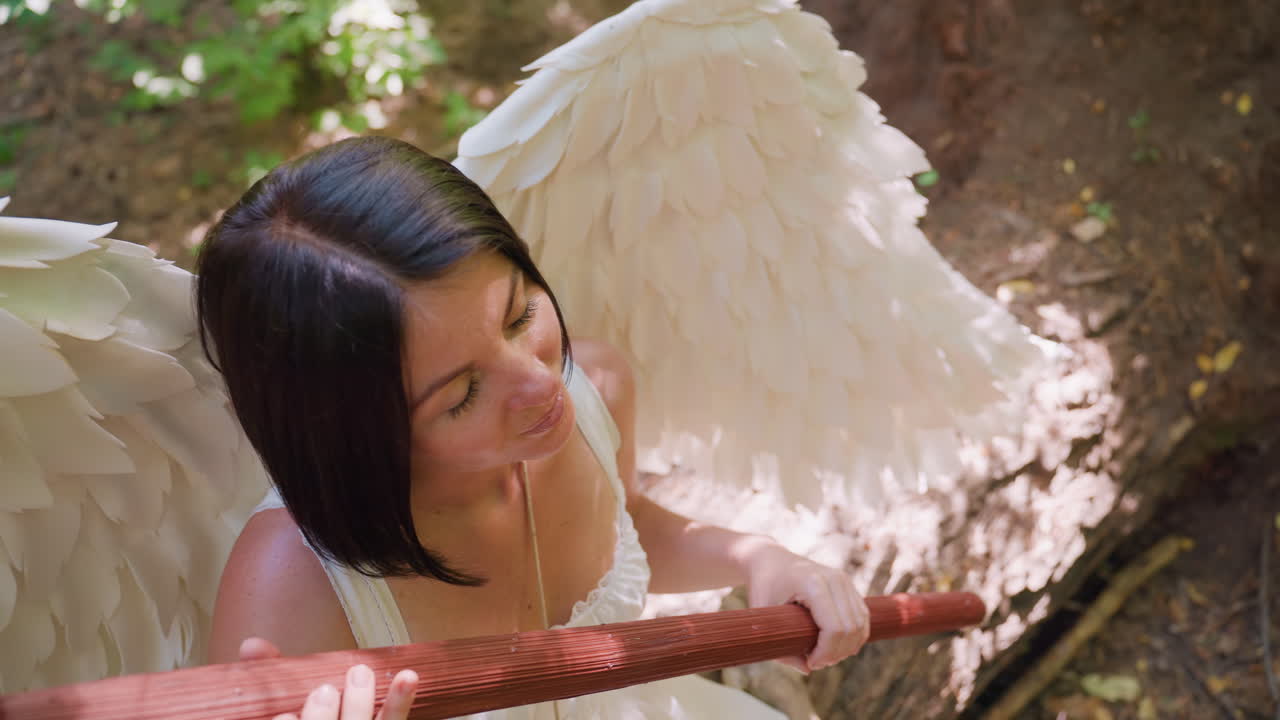 Aerial view of forest guardian with white wings seated under tree shade, gently bringing wooden staff close to chest, immersed in sunlight and peace, expressing spiritual harmony