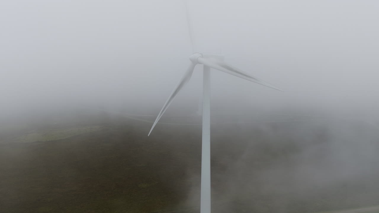 Drone shot moving through very heavy mist showing a wind turbine in West Yorkshire on a grey, misty and cold morning overlooking hills and fields