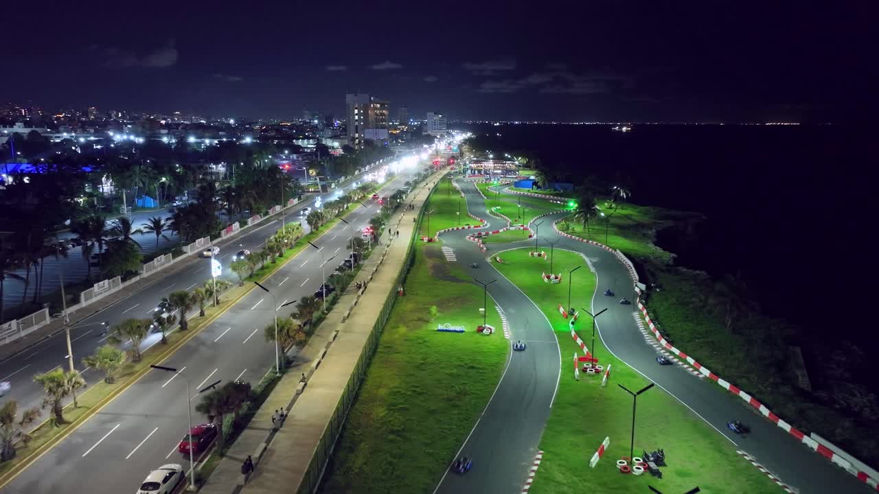 Aerial drone view of an illuminated go-kart track alongside the busy Malecón highway at night. City traffic flows past the racing circuit on the Caribbean coast of Santo Domingo, Dominican Republic