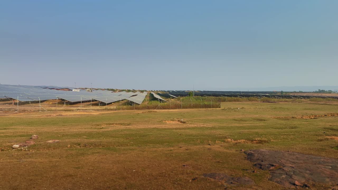 A wide panning shot moves across dry open land as rows of blue solar panels slowly enter the frame, revealing a sprawling solar farm stretching toward the horizon