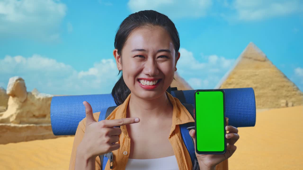 Close Up Of Asian Female Hiker With Mountaineering Backpack Smiling And Pointing To Mock Up Green Screen Smartphone While Traveling In Pyramid Of Giza