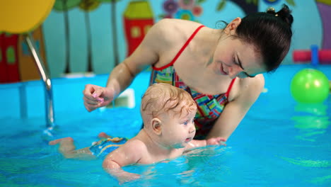female coach holding a chubby kid on her hand in the swimming pool. little infant splashing hands energetically.