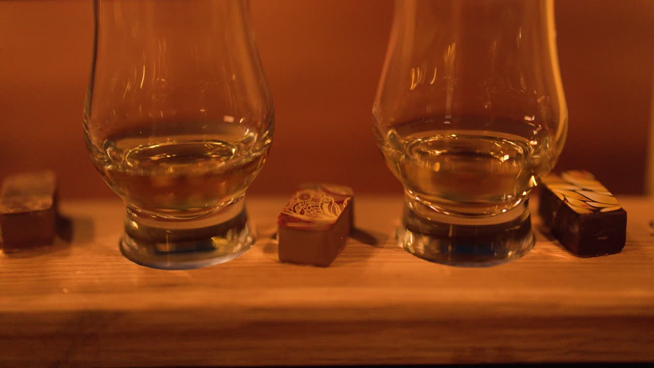 Close-up panning shot of whisky glasses and chocolates on wooden board, warm ambient lighting