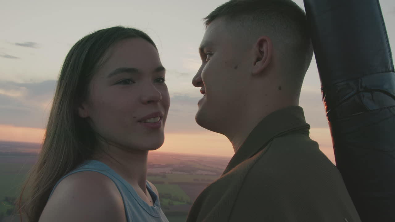 close up couple admiring vibrant sunset from hot air balloon basket while gazing into each other eyes surrounded by pastel sky and distant paraglider drifting above endless green fields at dusk