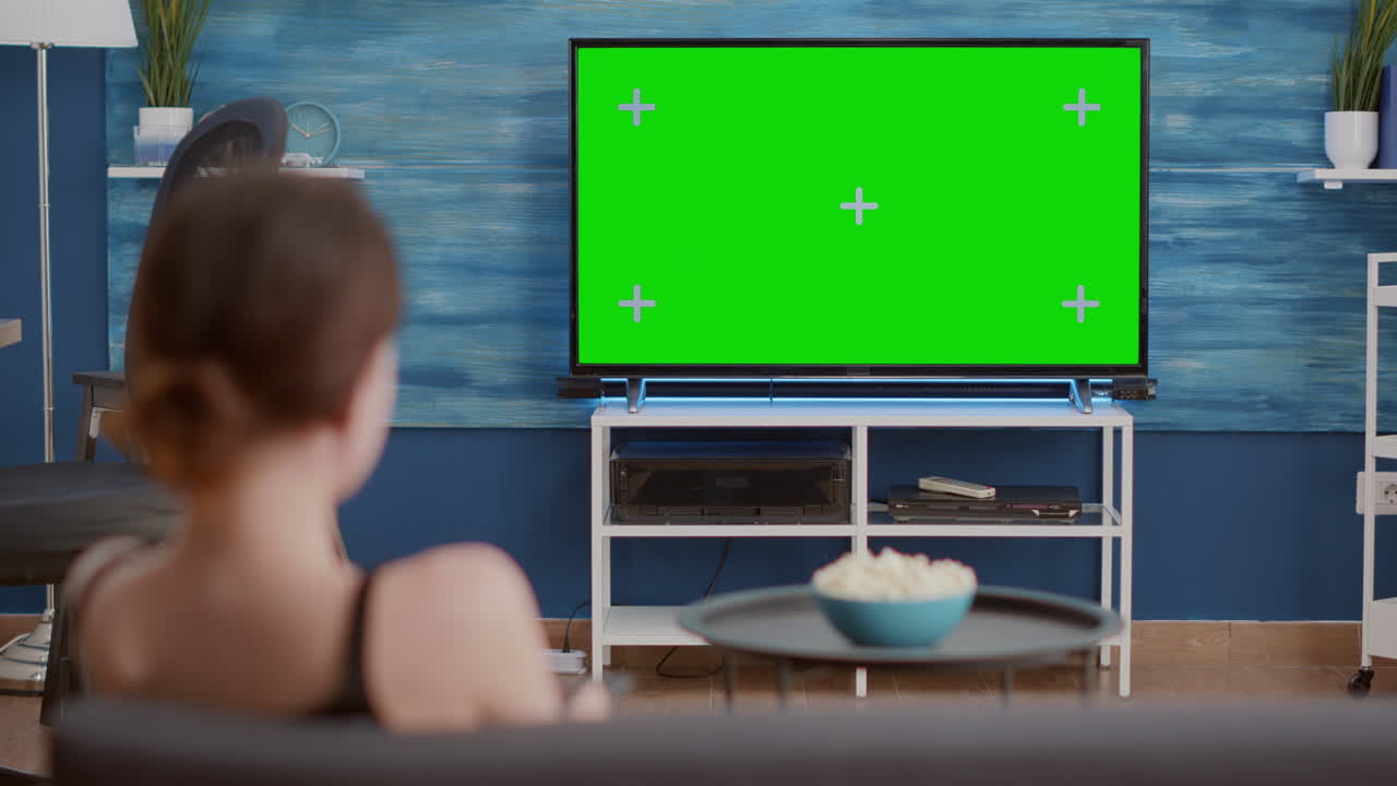 Static tripod shot of young woman switching channels while looking at green screen on tv