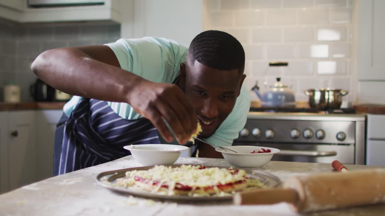 African american man making pizza in kitchen sprinkling grated cheese