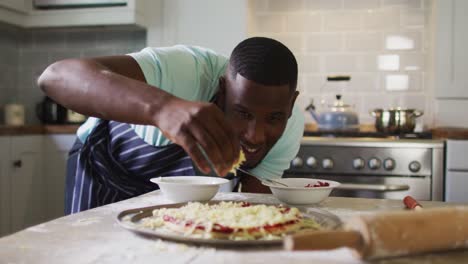 African-american-man-making-pizza-in-kitchen-sprinkling-grated-cheese