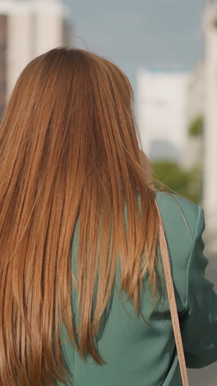 Woman with long loose red hair talks on phone walking along car parking in big city. Businesswoman conducts business remotely against blurry buildings backside view