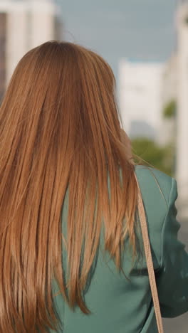 woman with long loose red hair talks on phone walking along car parking in big city. businesswoman conducts business remotely against blurry buildings backside view