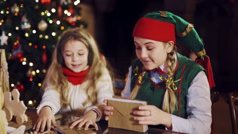 a funny elf is sitting at the table with a little girl and showing how to tie a stack of envelopes with wishes using a rope in the residence of santa claus. blurred background