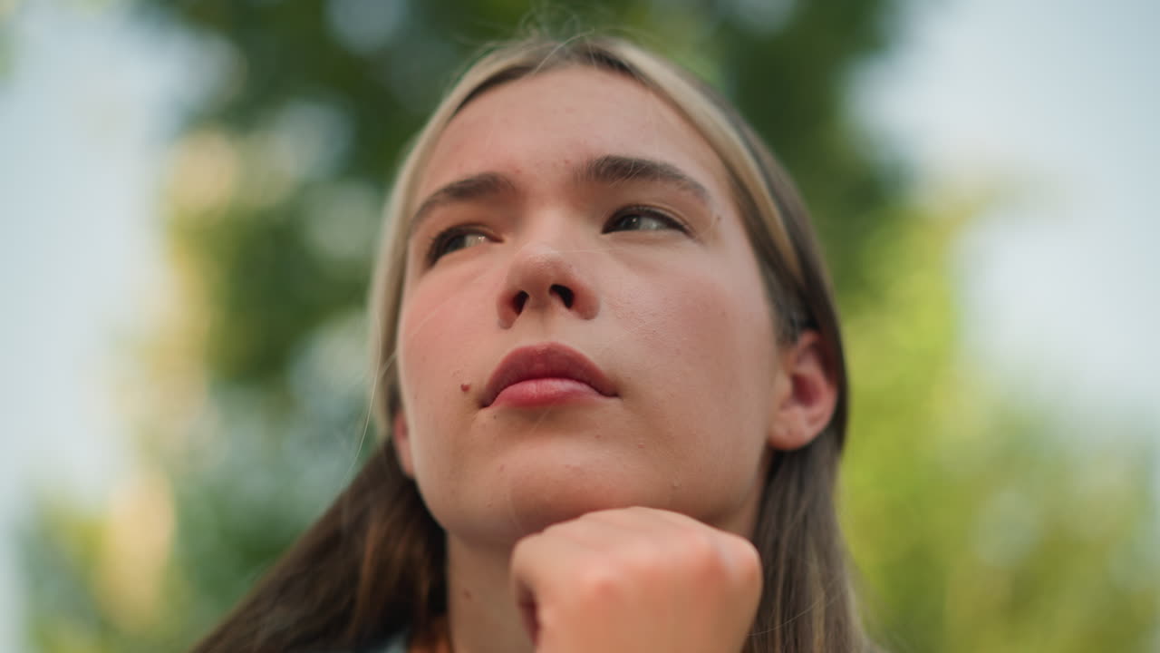 primer plano de una mujer joven con la mano debajo de la barbilla, mirando pensativa mientras mira a la distancia, expresando un pensamiento profundo vegetación borrosa en el fondo, la luz del sol ilumina suavemente su rostro