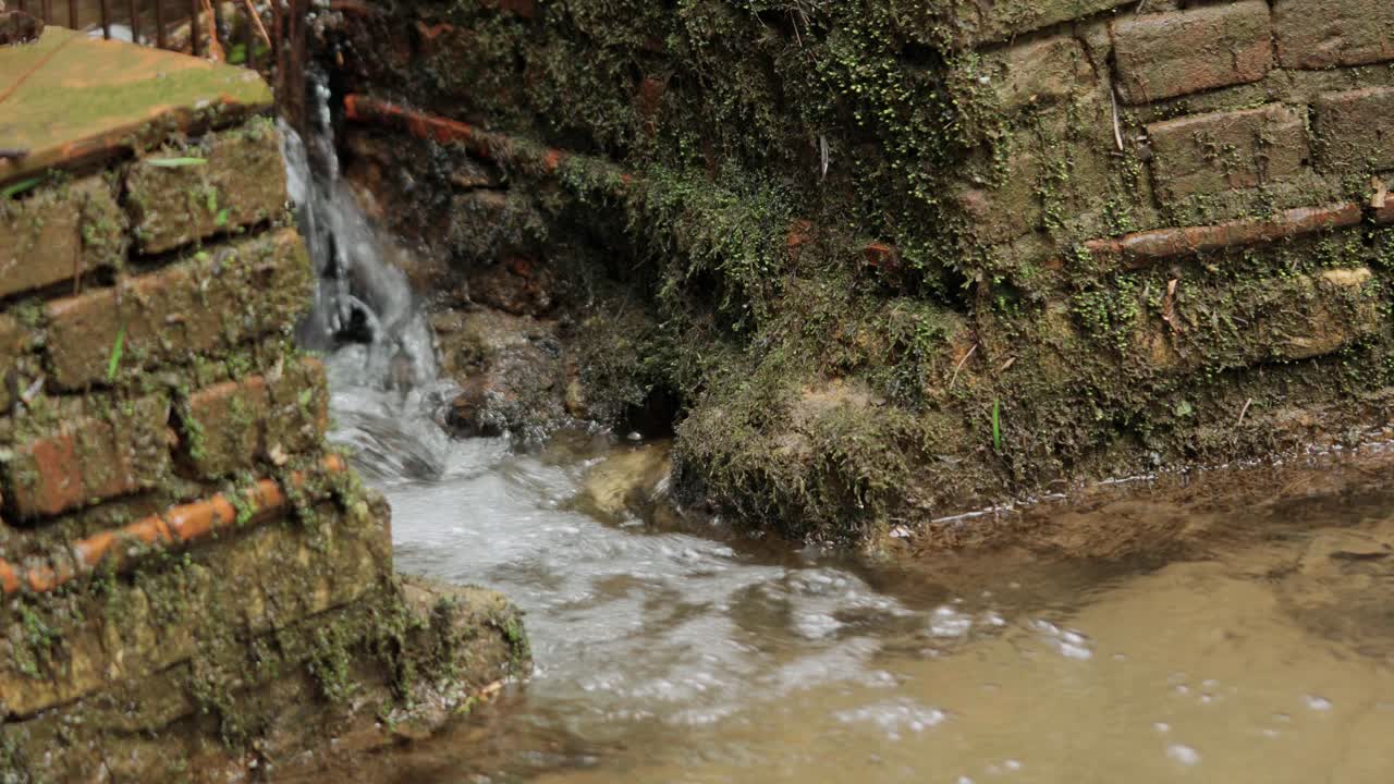 Fresh water flowing continuously from a rustic stone and brick outlet into a tranquil small lake in a park setting