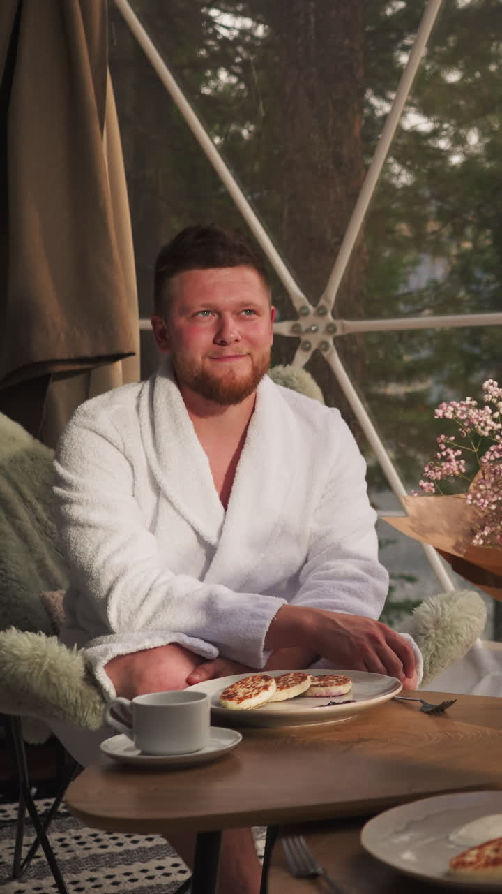 Man sits at breakfast table opposite woman. Young husband and wife have sweet pancakes for breakfast near panoramic window in glamping tent house