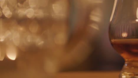 a hand reaches for a whisky glass on a wooden table, surrounded by warm candlelight and blurred background, creating a cozy, intimate atmosphere