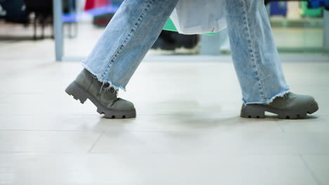 vista lateral de la pierna de alguien con pantalones vaqueros y botas llevando una bolsa de compras en la mano izquierda caminando en el centro comercial, sombra en el piso de azulejos, ropa borrosa visible a través del vidrio en el fondo
