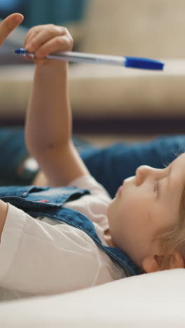 close-up little pretty daughter student in blue denim sundress, with light hair with pigtails, child lying on floor draws ballpoint pen in book, then her dad, thoughtfully looking to side forward