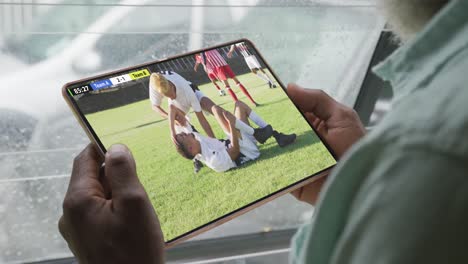 African-american-man-using-tablet-with-diverse-male-soccer-players-playing-match-on-screen