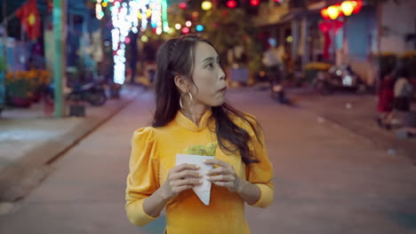 slow motion of vietnamese woman in traditional clothing walking and eating local pizza in hoi an street at night