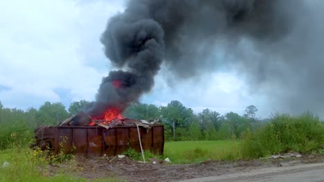 thick dark smoke billowing from a burning dumpster full of garbage on rural farmland