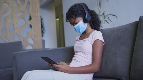 chica afroamericana con máscara facial usando una tableta digital mientras está sentada en el sofá en el hospital