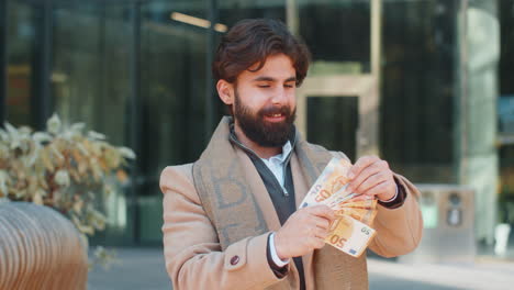 successful rich happy young indian man holding euro bill money cash fan on downtown city street