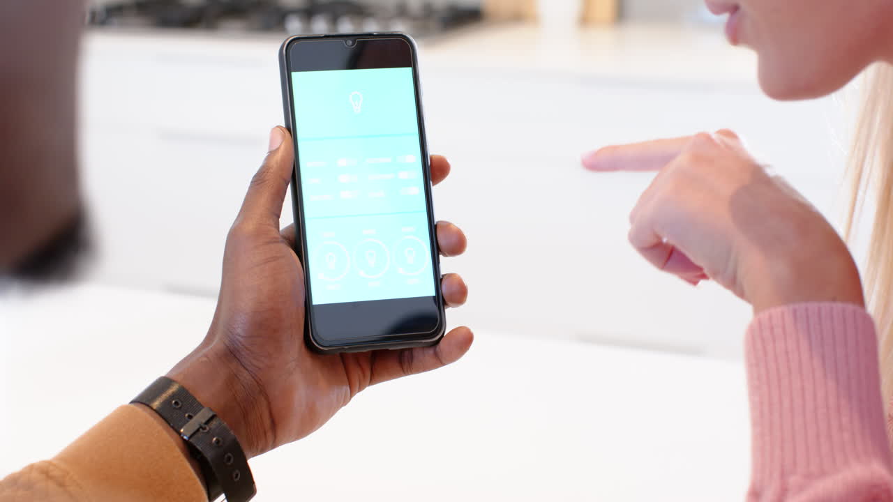diverse couple using smartphone app to control smart home lighting at kitchen table