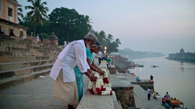 Couple de personnes âgées s'approchant, en kurta et sari, déposant des guirlandes rouges et blanches sur le mur d'un ghat en guise d'offrande