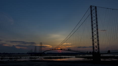 Evening-timelapse-of-Chandani-Dodhara-Suspension-Bridge-with-the-sun-setting-in-the-background-and-people-crossing-the-bridge-over-Karnali-River,-West-Nepal